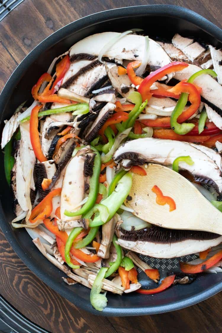 Frying pan with mushrooms and peppers being mixed by wooden spoon