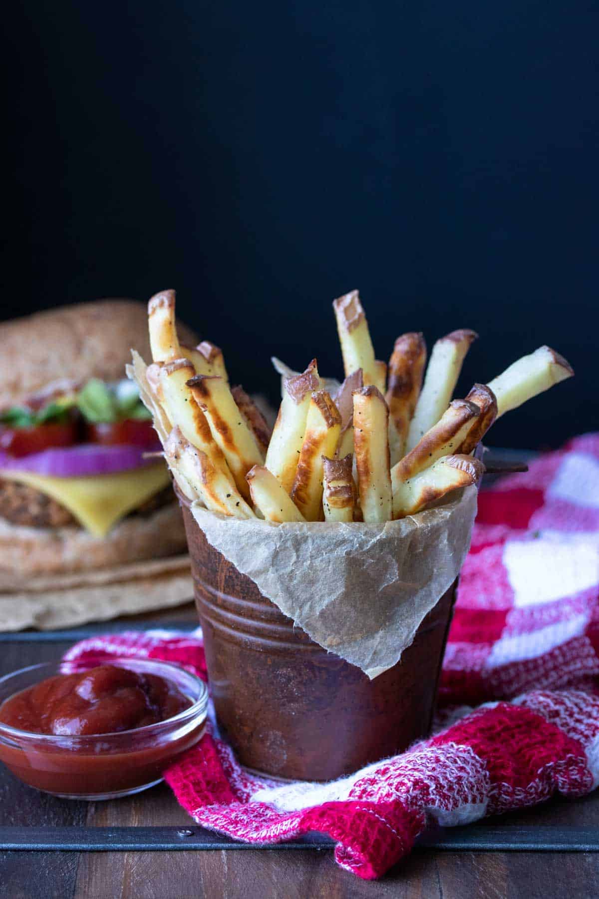 Brown pail on a red checkered towel filled with crispy french fries next to ketchup.