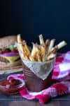 Baked french fries in a brown pail in front of a burger on a red checkered towel.