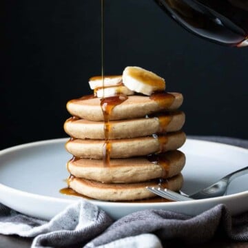 Maple syrup being poured onto a stack of pancakes on a grey plate