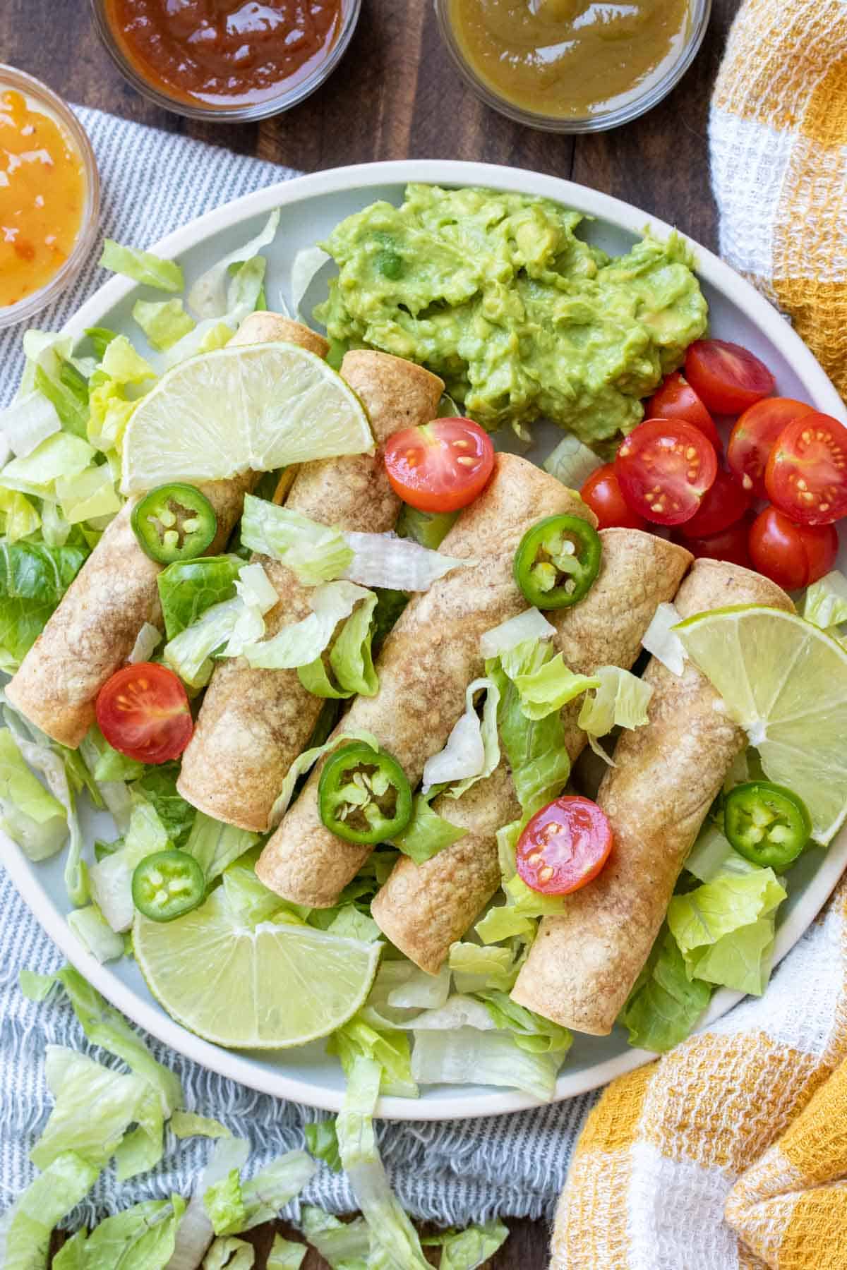 White plate with a row of taquitos surrounded by lettuce, tomatoes and guacamole