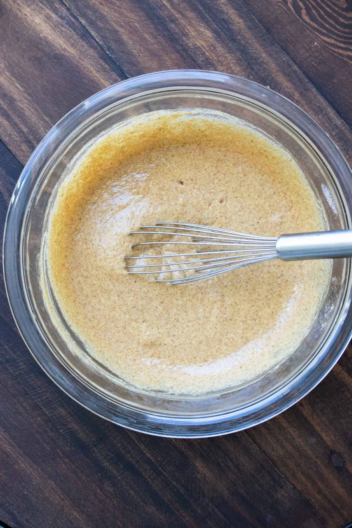 Whisk mixing tan cupcake batter in a glass bowl.