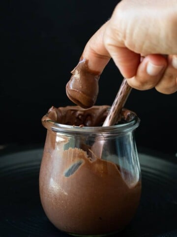 Finger taking a scoop of chocolate frosting from a glass jar