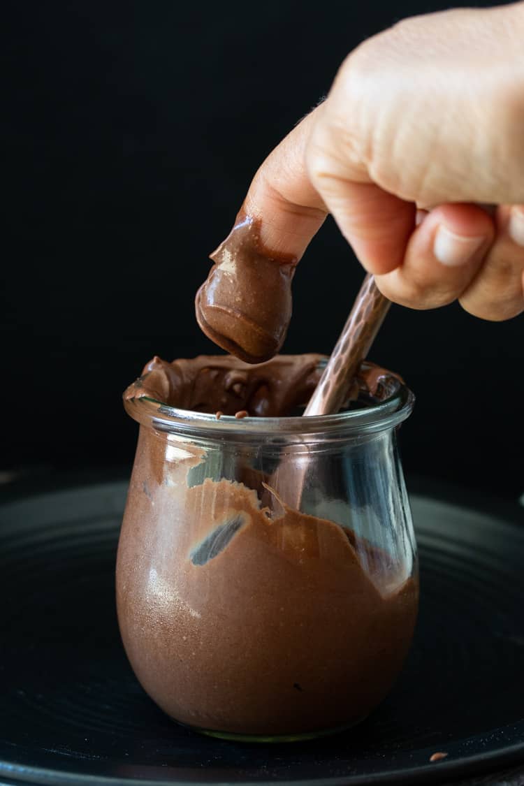 Finger taking a scoop of chocolate frosting from a glass jar
