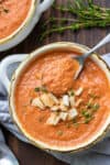 A metal spoon taking a bite of tomato soup from a white soup bowl with handles topped with crackers and thyme.