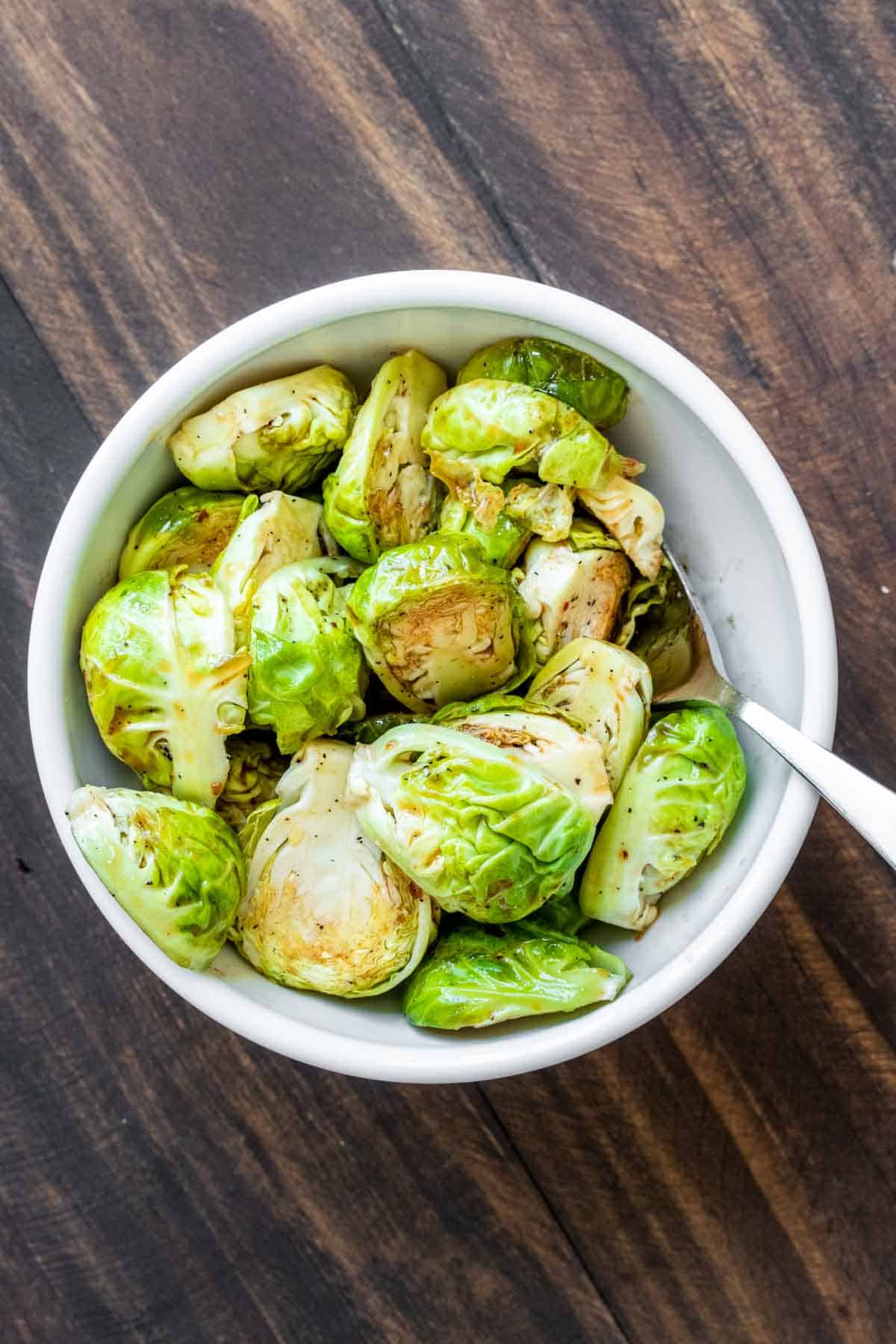 Spoon mixing Brussels sprouts with maple syrup and seasoning in a white bumpy bowl.