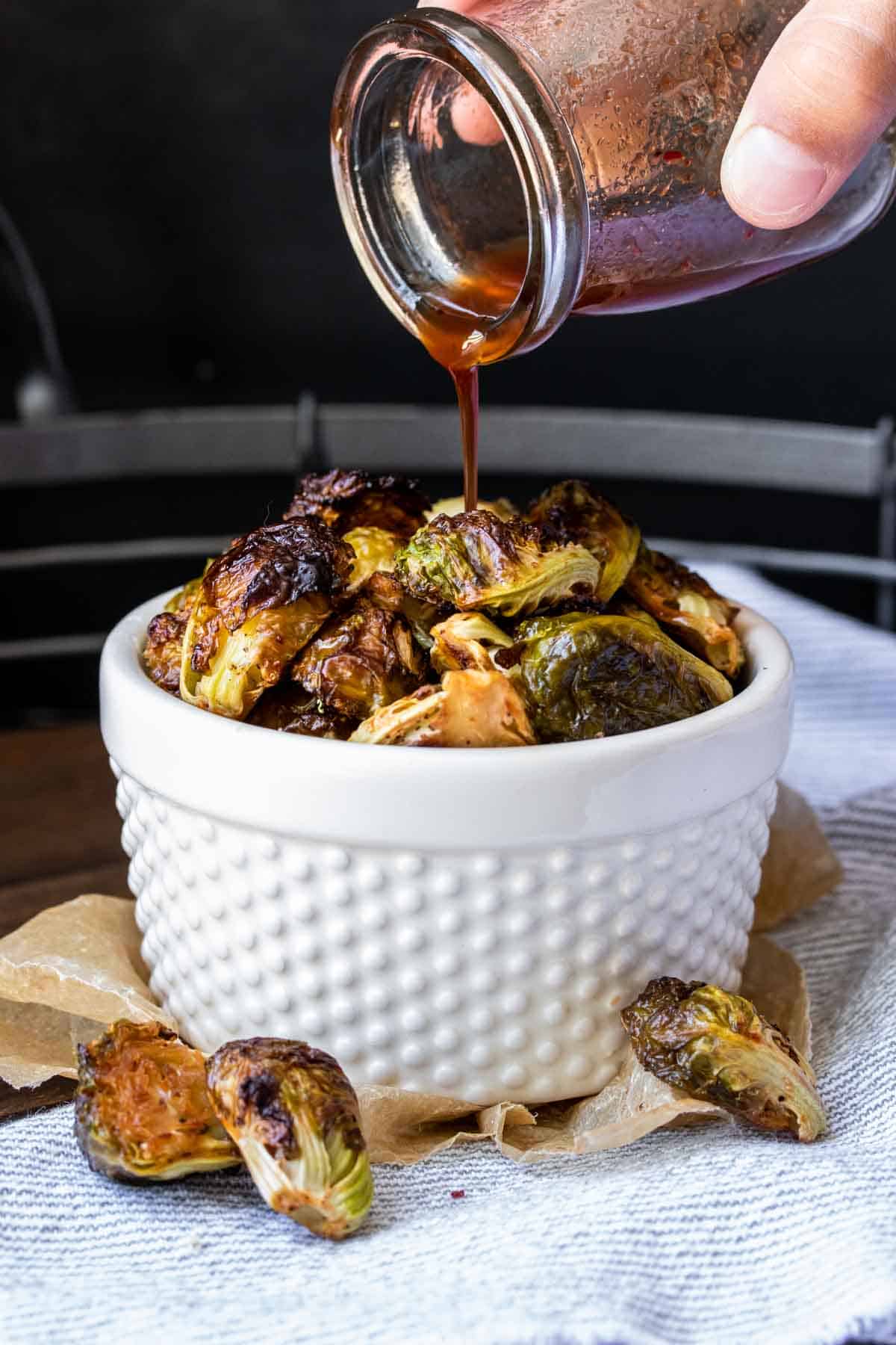 Hand pouring maple syrup sauce over a bowl of roasted Brussels sprouts.