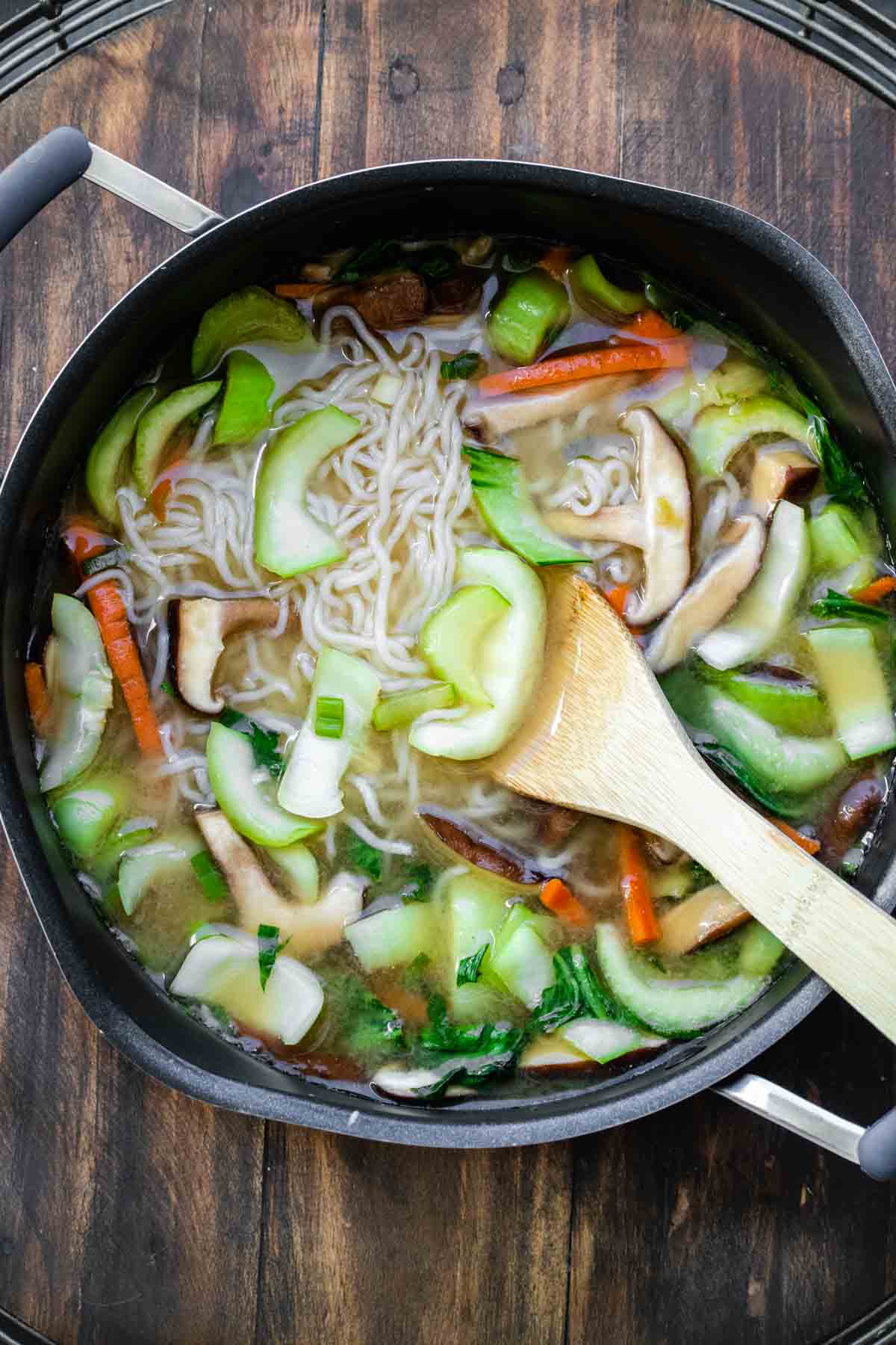 Veggies and noodles in broth being stirred in a pot.