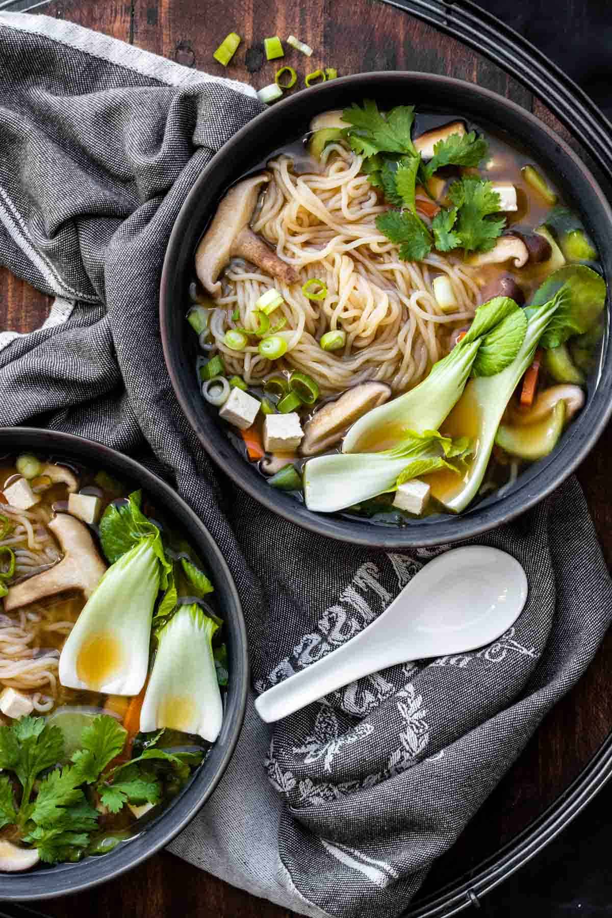 Two bowls of veggies and noodle soup on a wooden table.