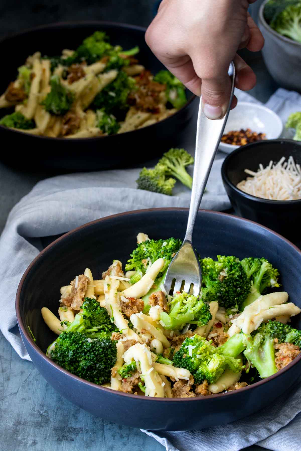 Someone taking a bite of cavatelli and broccoli with a fork from a blue bowl in front of another bowl and ingredients