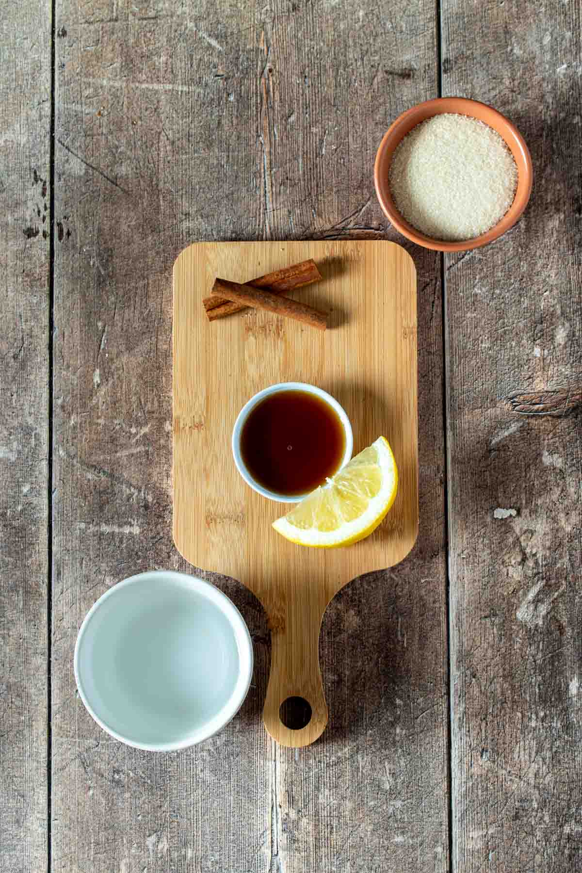 Top view of a wooden cutting board with a cinnamon stick, maple syrup and lemon wedge on it and sugar and water next to it. 