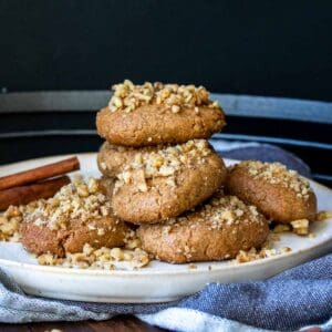 A pile of walnut covered cookies on a white plate with a cinnamon stick in the back.