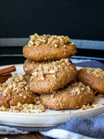 A pile of walnut covered cookies on a white plate with a cinnamon stick in the back.