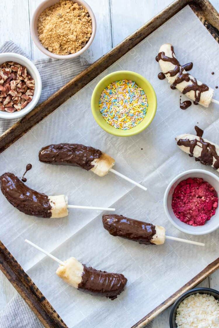 Bananas on sticks covered in chocolate on a cookie sheet with toppings in bowls around them