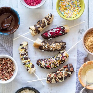 A group of bananas covered in chocolate and toppings sitting on a piece of parchment paper on a white wood surface