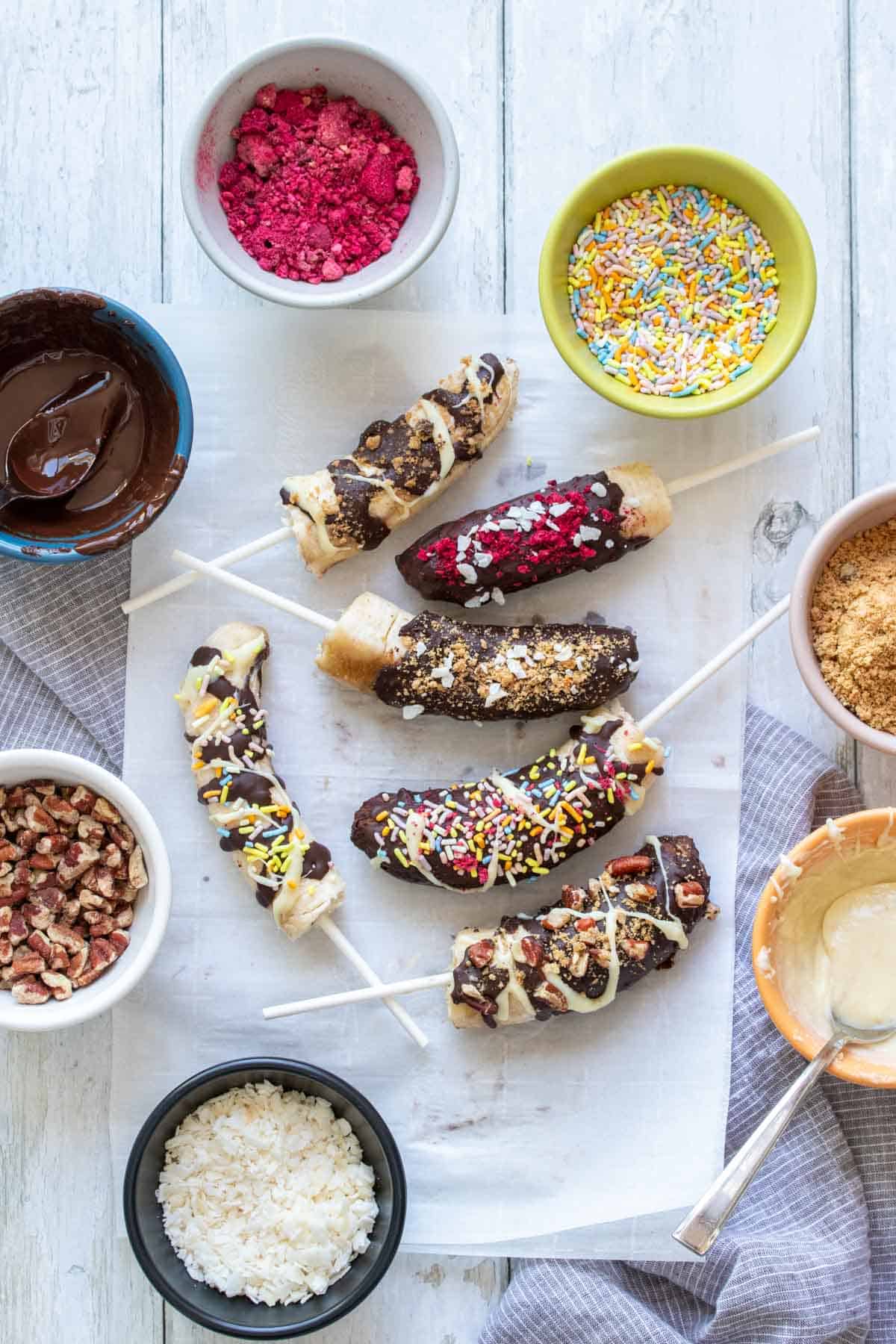 A group of bananas covered in chocolate and toppings sitting on a piece of parchment paper on a white wood surface