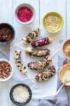 Chocolate covered bananas on sticks sprinkled with a variety of toppings and bowls of toppings around them