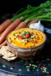 A carrot hummus in a brown bowl next to crackers, carrots and garlic on a dark surface.