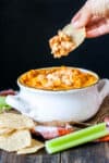Baked buffalo chicken dip in a white bowl with handles and a chip with dip on it coming out of the bowl.