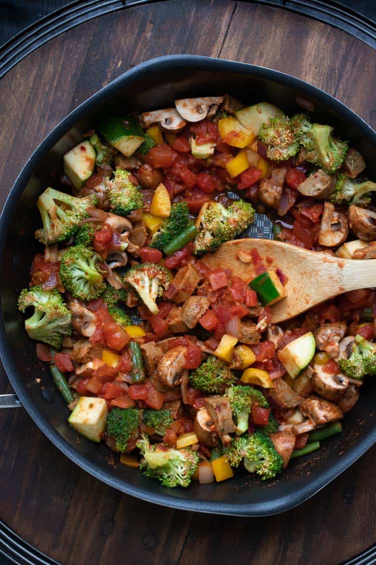 Wooden spoon mixing sautéed veggies and tomatoes in a pan