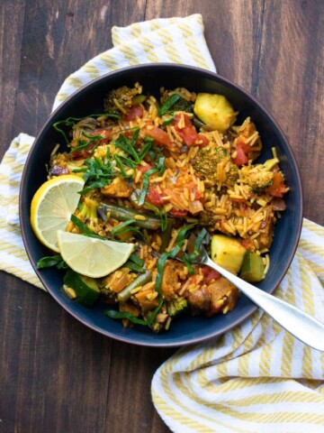 Black bowl of veggie paella topped with lemon slices and strips of greens