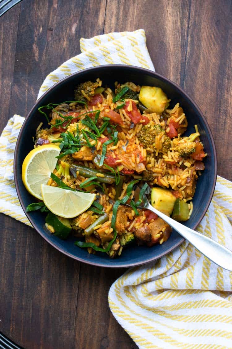 Black bowl of veggie paella topped with lemon slices and strips of greens