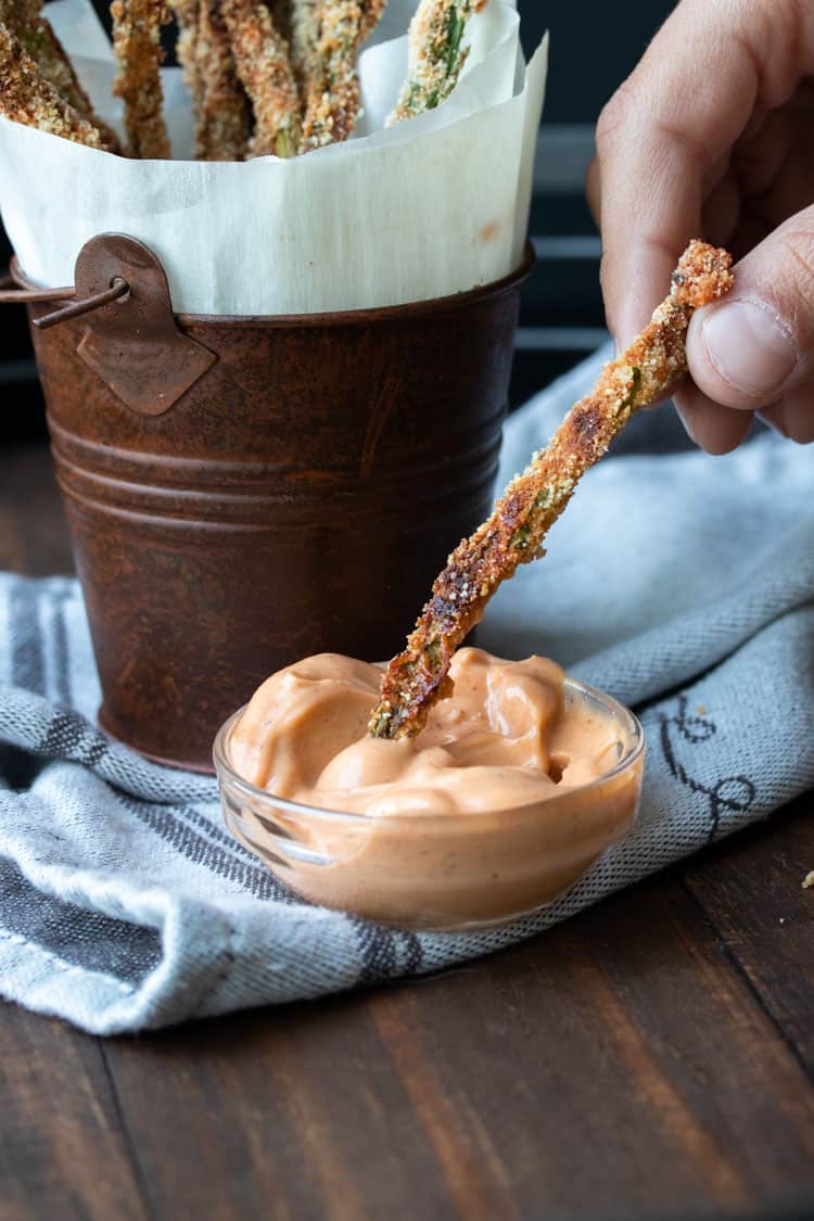 Hand dipping a crispy breaded asparagus fry into creamy pink sauce