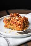 A white plate with a piece of vegetable lasagna with red sauce on it sitting on a grey towel.