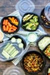 A group of black containers with plastic lids filled with veggies sitting on a blue striped towel on a wooden surface.