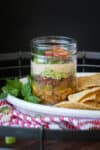 A white plate with chips and a glass jar layered with a Mexican 7 layer dip.