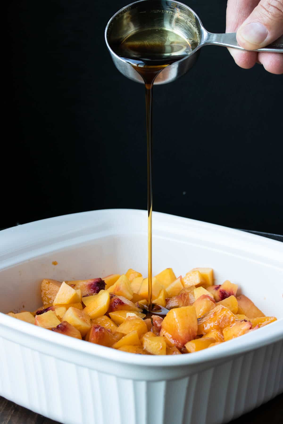 Maple syrup being poured into a white baking dish filled with peaches.