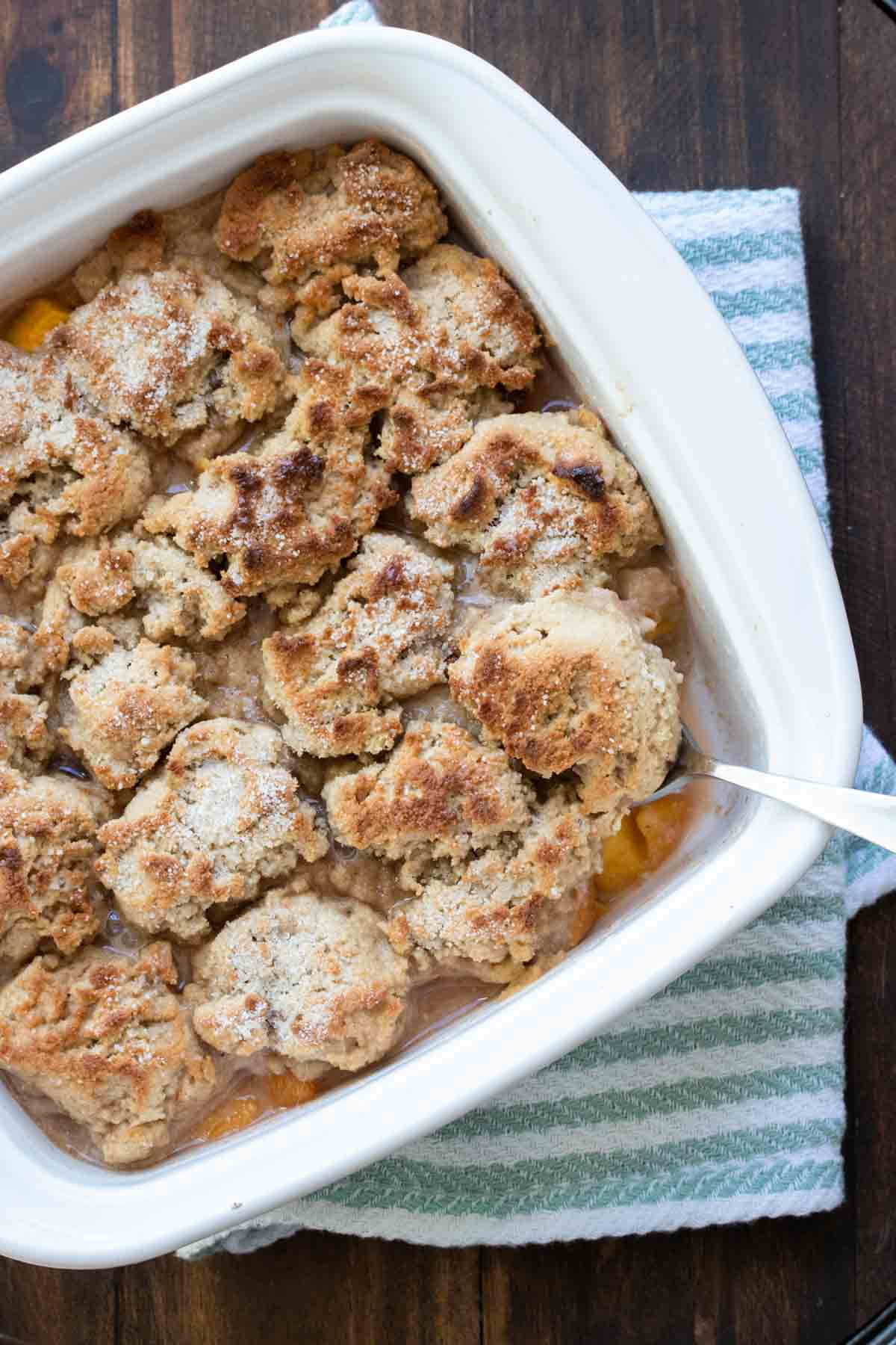 Spoon taking a scoop out of a white baking dish with baked peach cobbler.