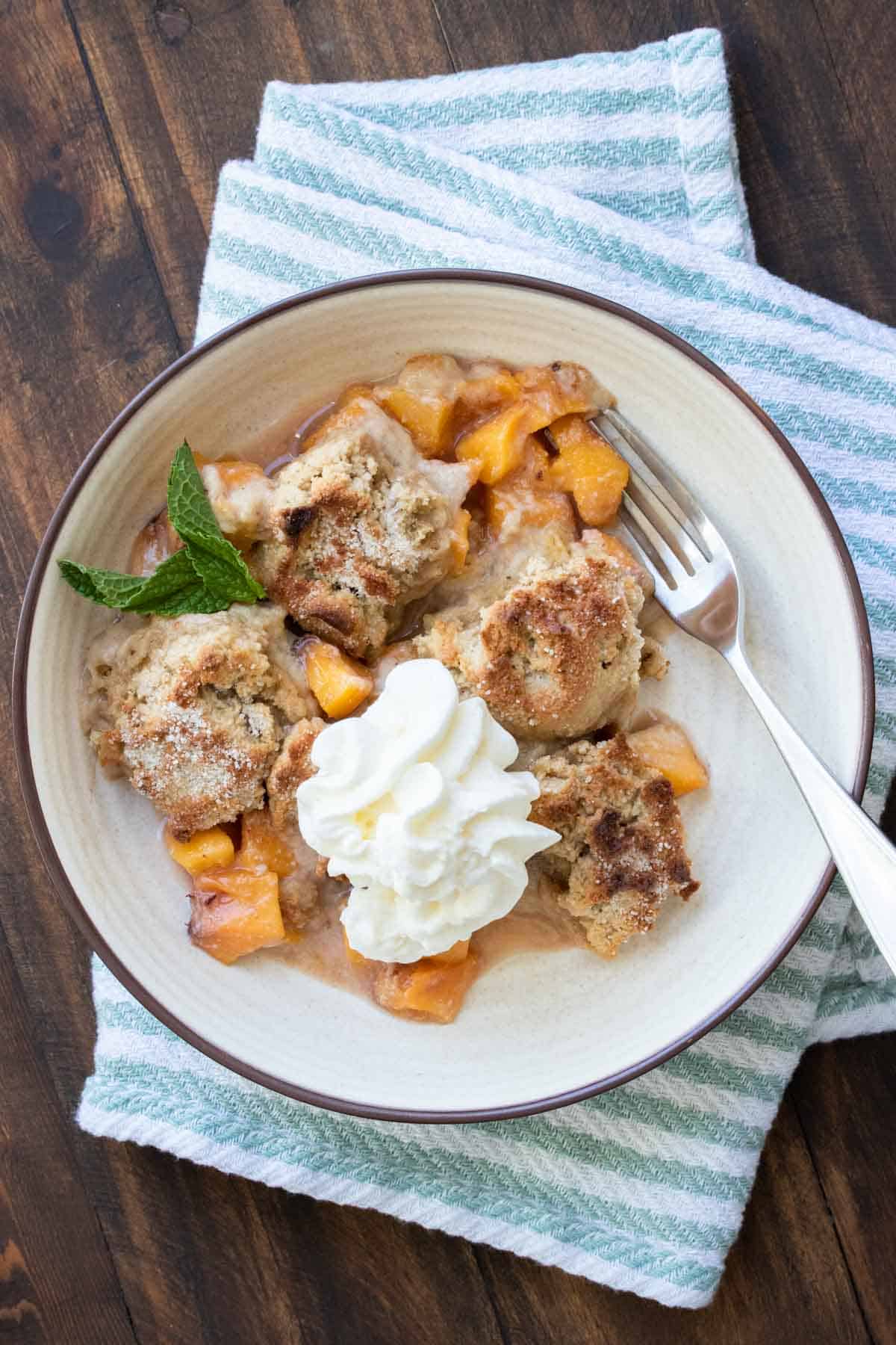 Peach cobbler with a fork and topped with whipped cream and a mint leaf in a cream bowl.