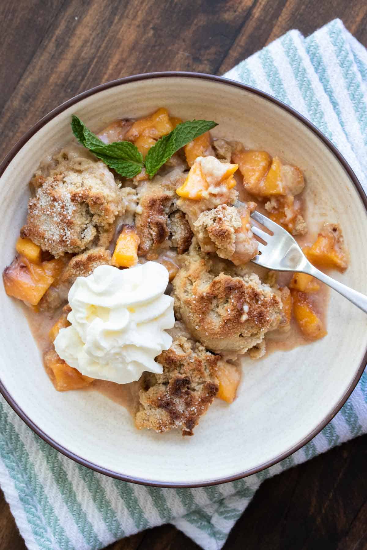 Fork taking a bite of peach cobbler from a cream bowl