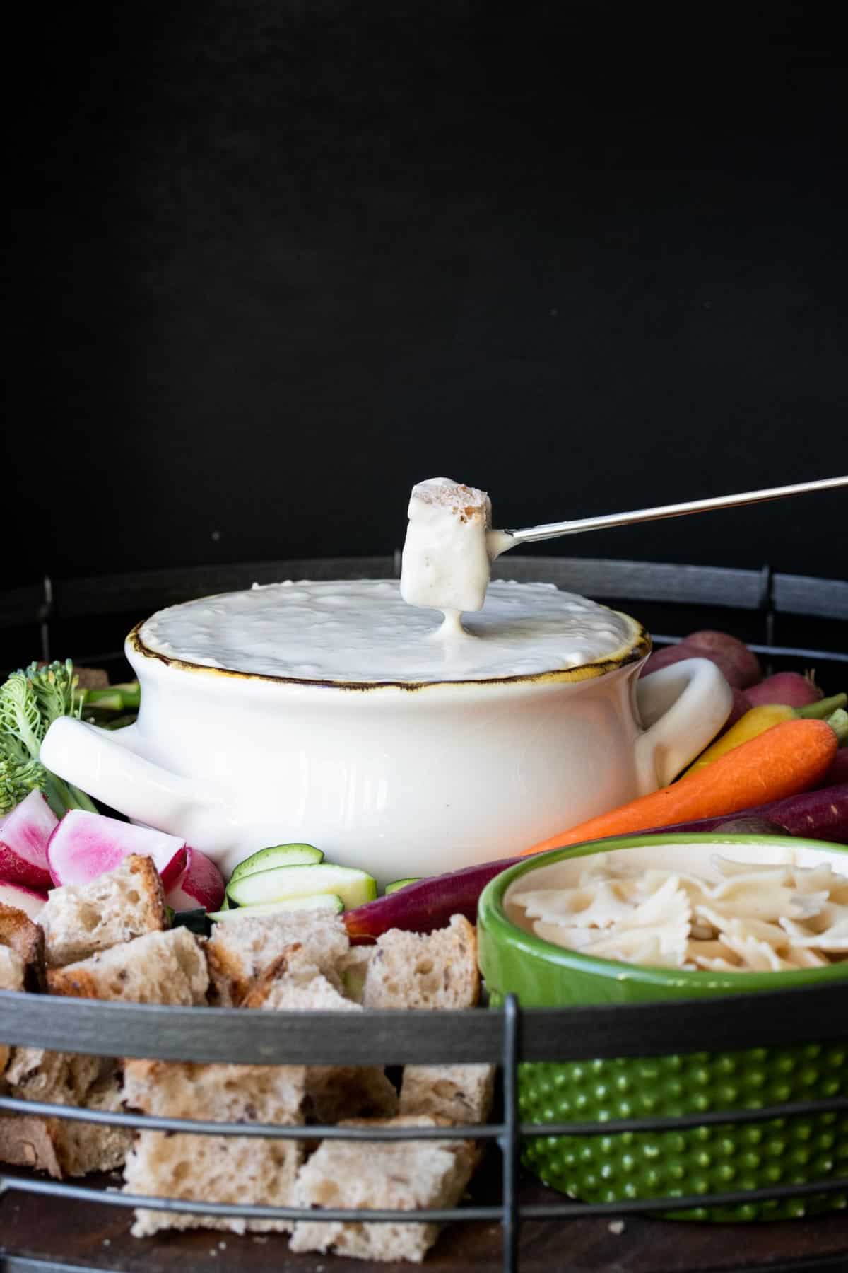 Piece of bread being dipped into a bowl of cheese fondue surrounded by veggies.