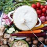 A bowl of cheese fondue with veggies and bread around it and a piece of bread dipping in.