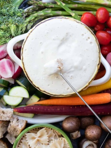 A bowl of cheese fondue with veggies and bread around it and a piece of bread dipping in.