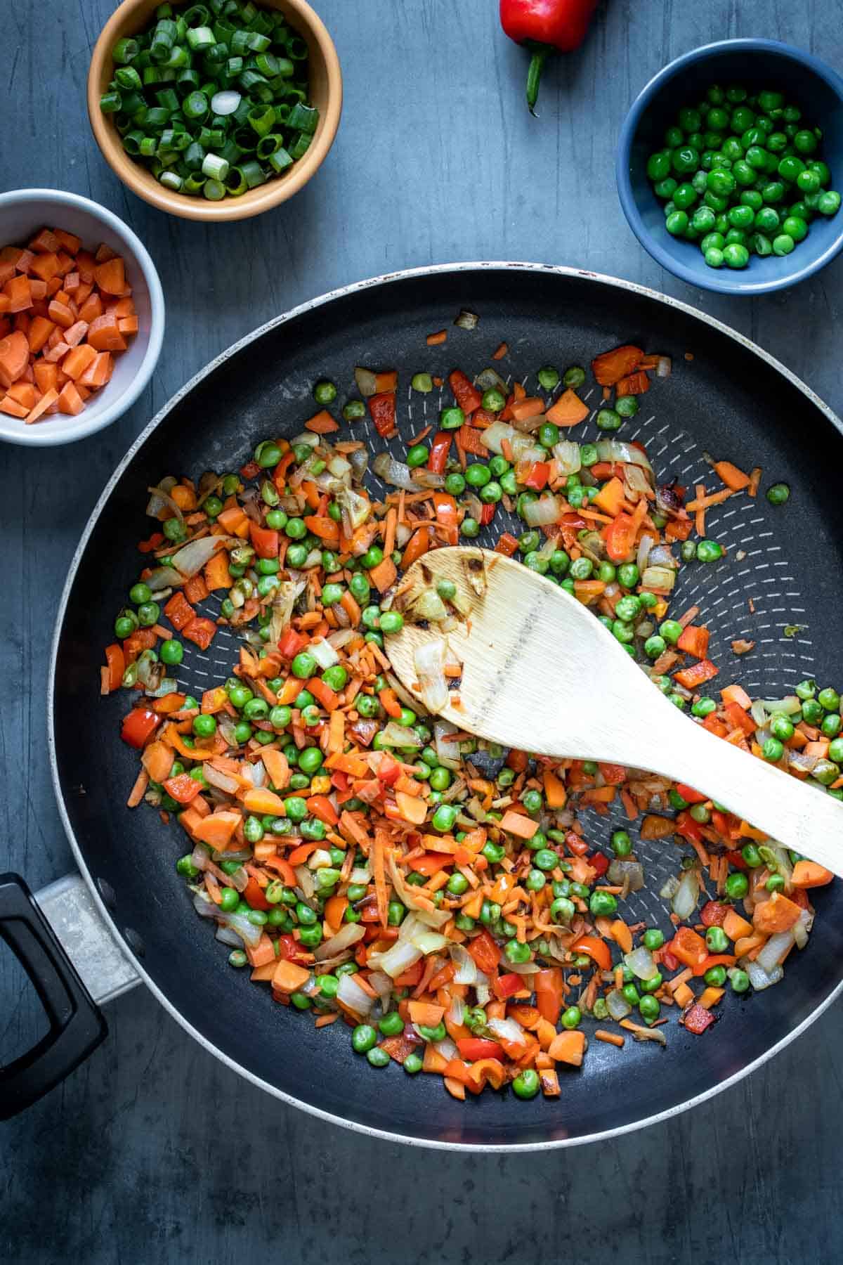 Wooden spoon mixing veggies cooking in a pan surrounded by bowls