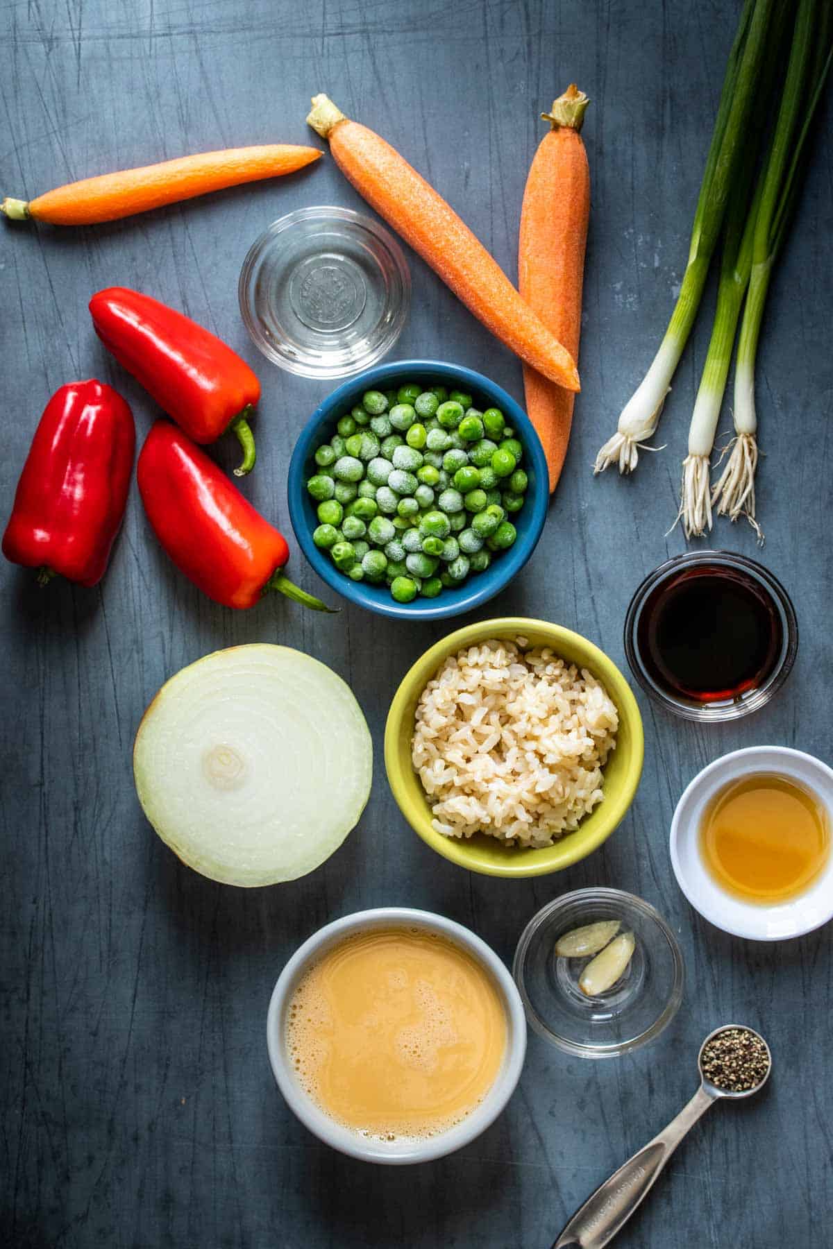 Bowls with ingredients next to veggies for a fried rice recipe on a dark surface