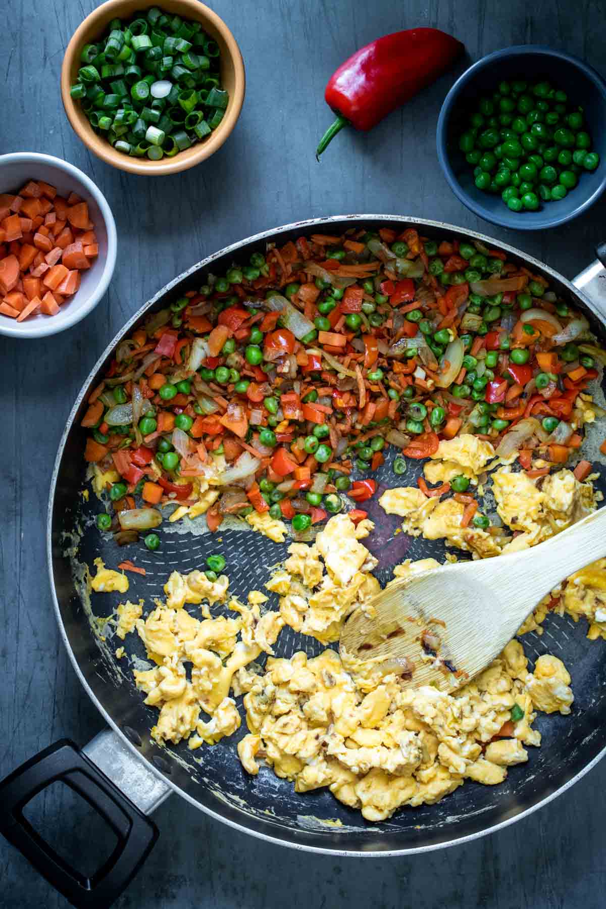 A wooden spoon mixing scrambled eggs in a pan next to chopped veggies with small bowls surrounding it