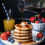 Maple syrup being poured over a stack of five pancakes on a white plate