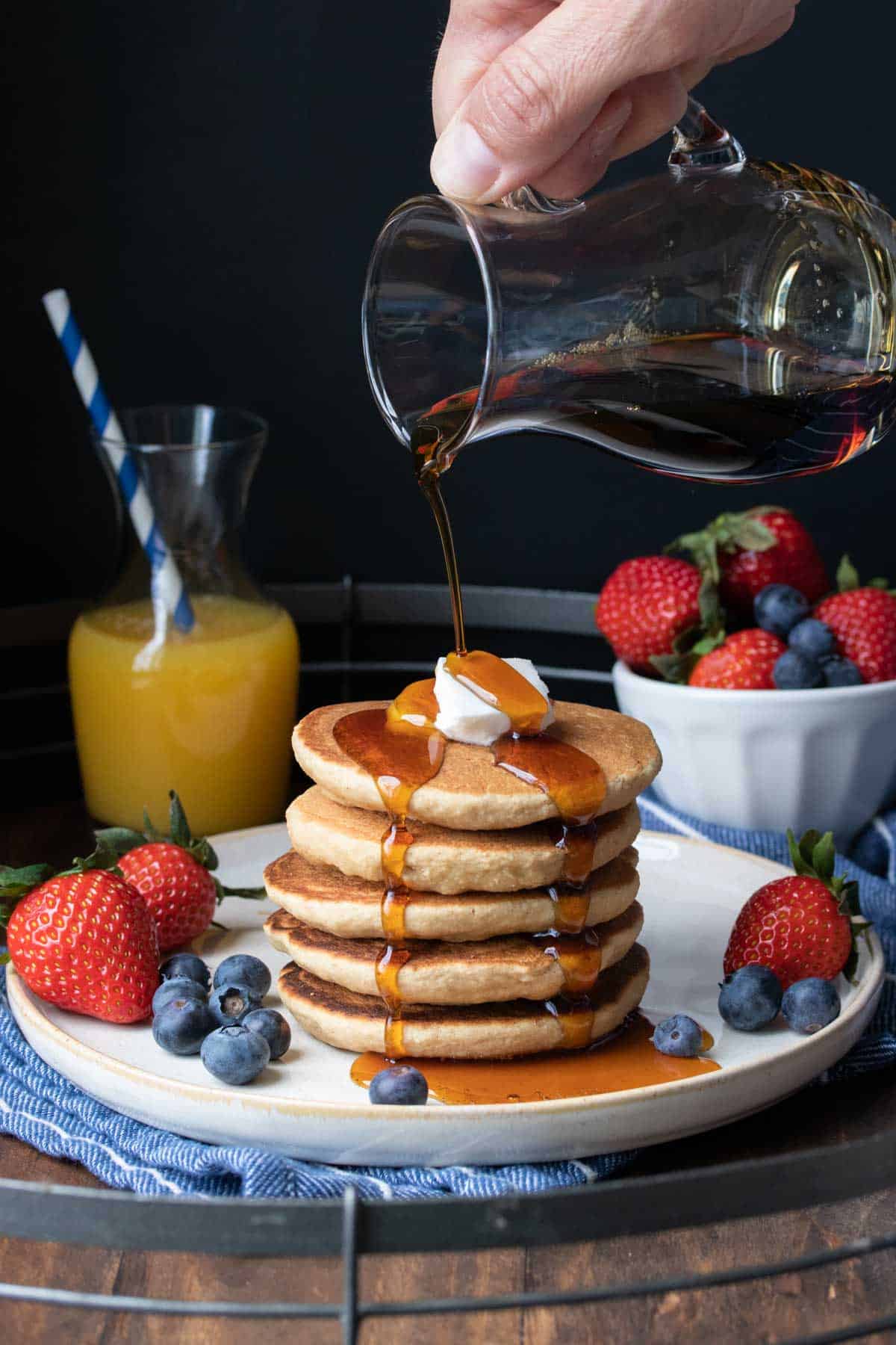 Maple syrup being poured over a stack of five pancakes on a white plate