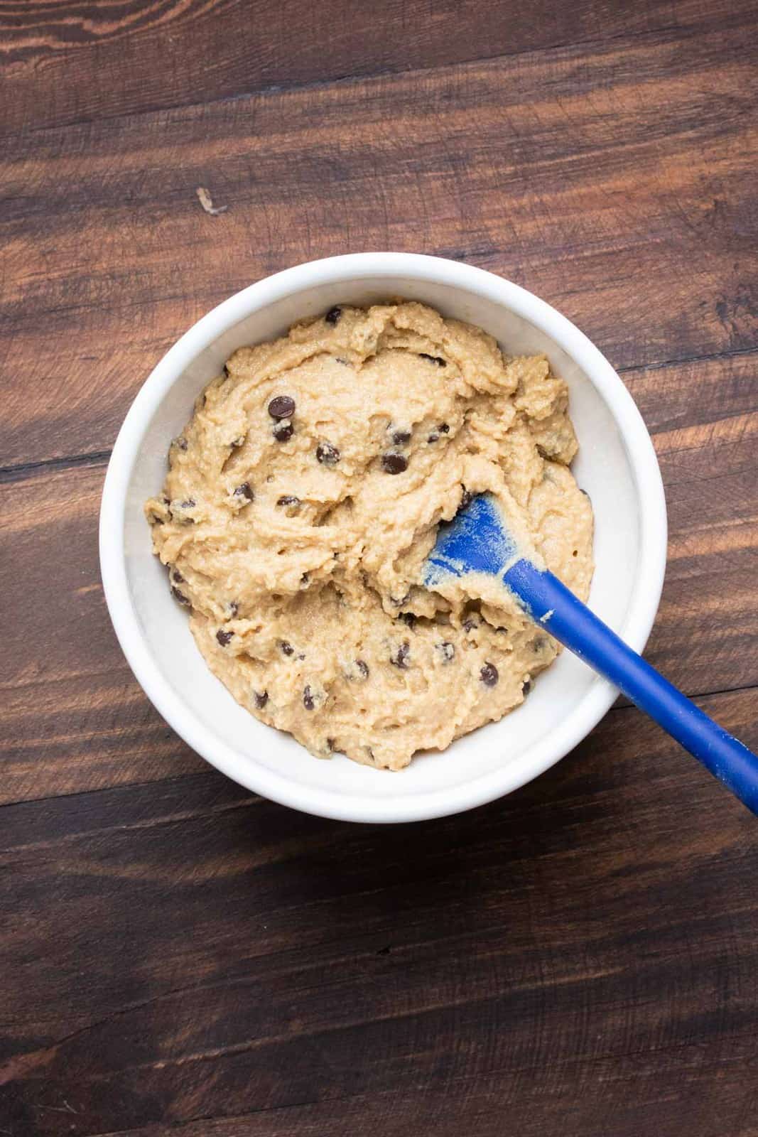 Blue spatula mixing chocolate chip cookie dough in a white bowl