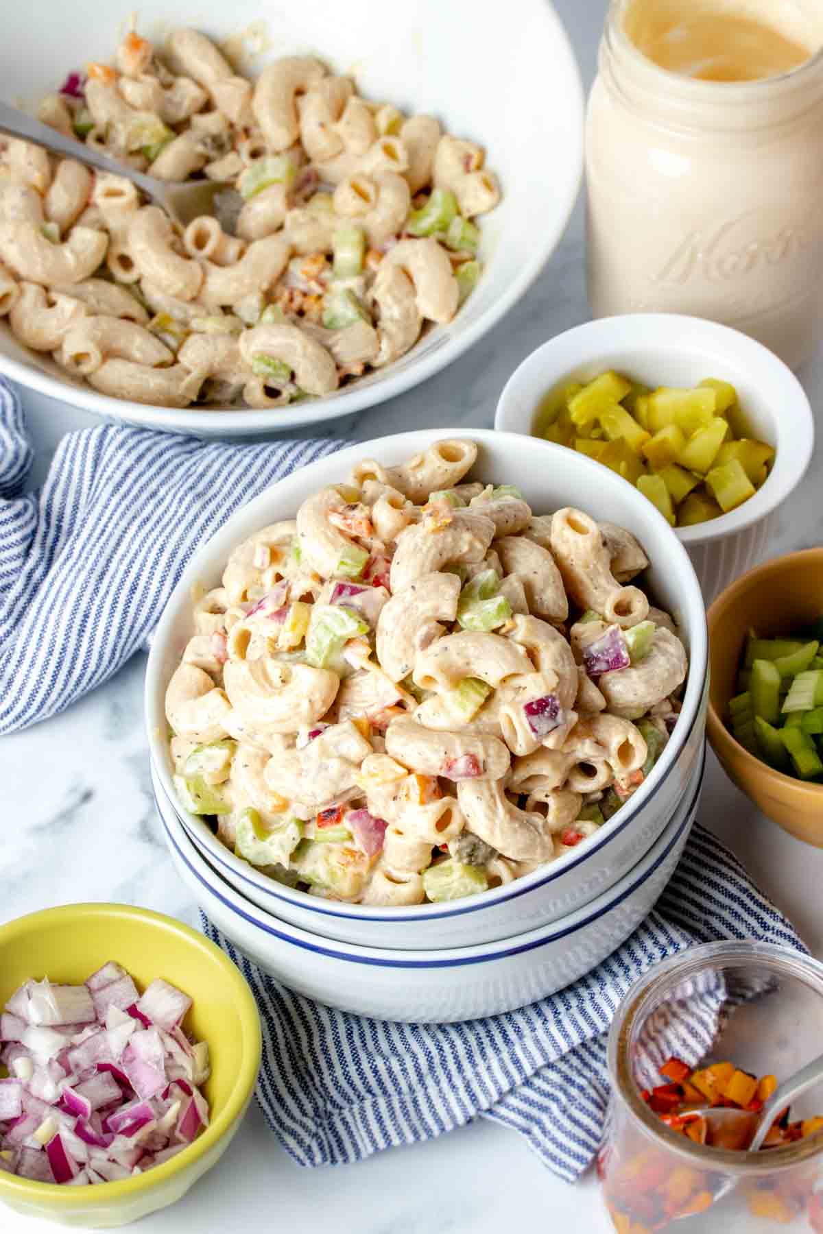 Top side view of two white bowls stacked and filled with creamy macaroni salad surrounded by ingredients in smaller bowls.