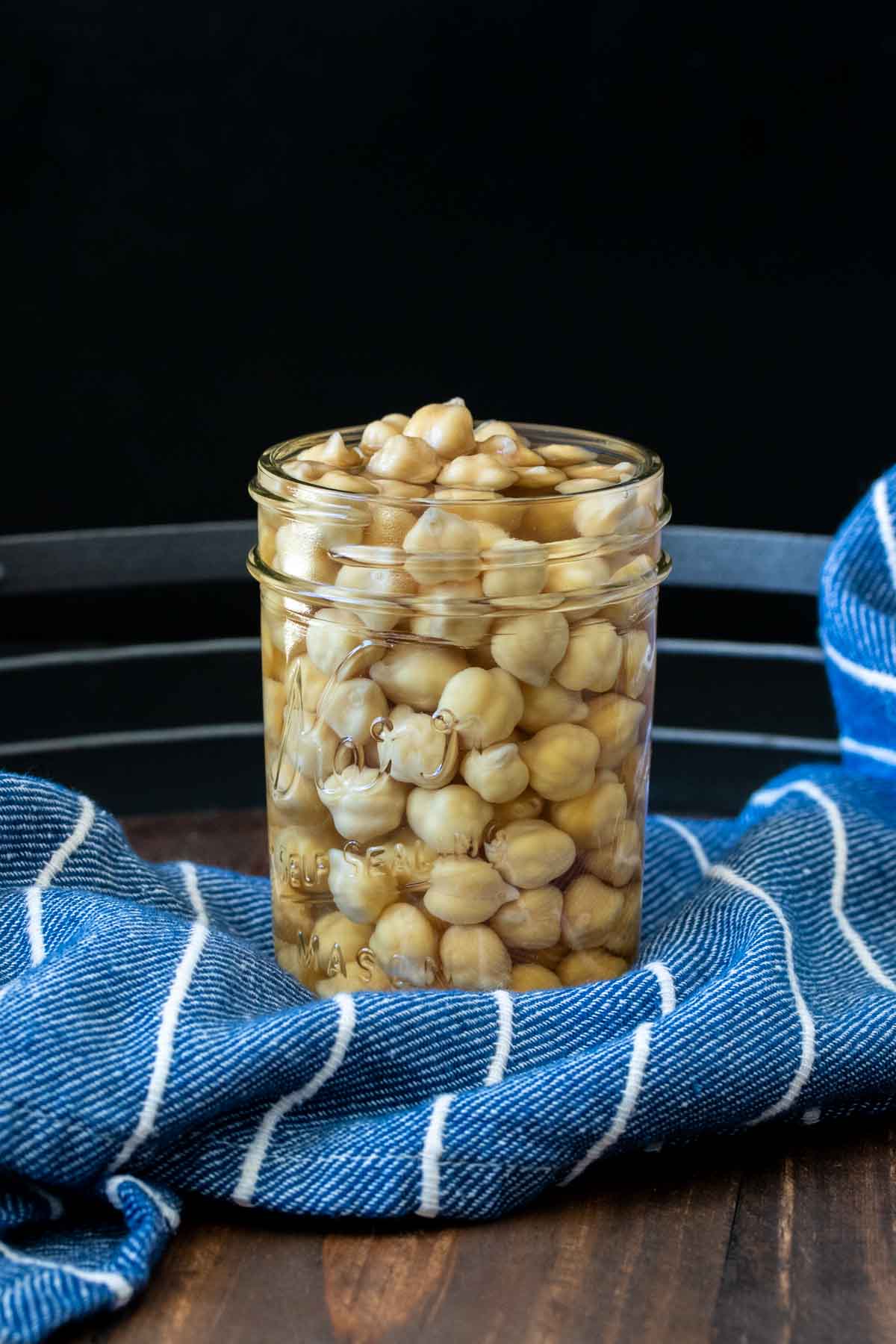 Glass jar filled with soaked dried chickpeas in water sitting on a blue striped towel.