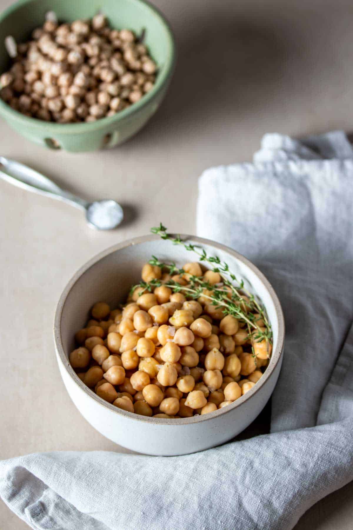 A white bowl filled with chickpeas and thyme sprigs on a tan surface in front of a green strainer with dry chickpeas.