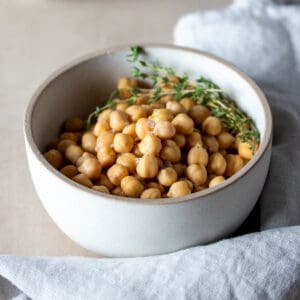 A white bowl surrounded by a towel with cooked chickpeas inside and sprigs of thyme on top.