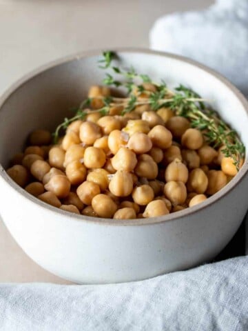 A white bowl surrounded by a towel with cooked chickpeas inside and sprigs of thyme on top.