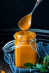 A metal spoon with buffalo sauce dripping from it into a glass jar on a blue towel.