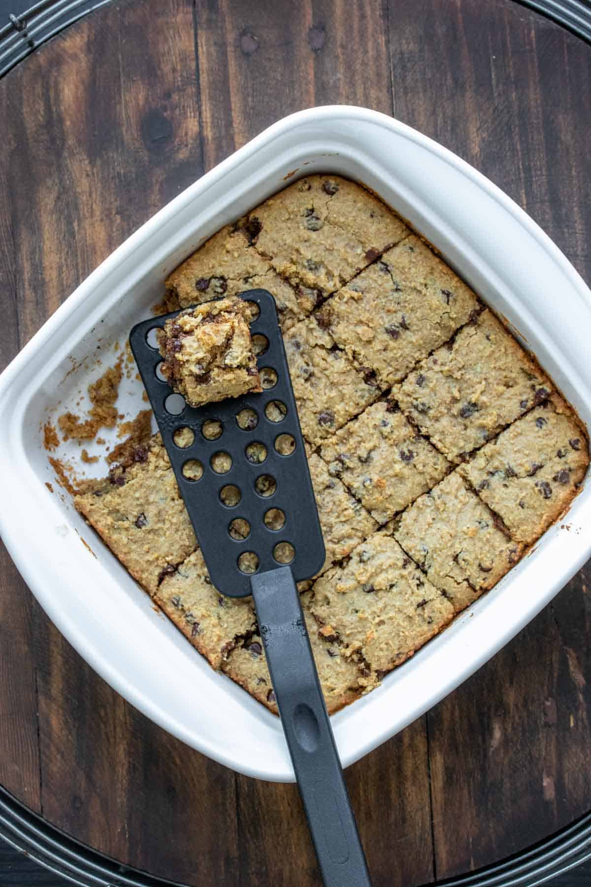 Black spatula taking a blondie piece from a white baking dish